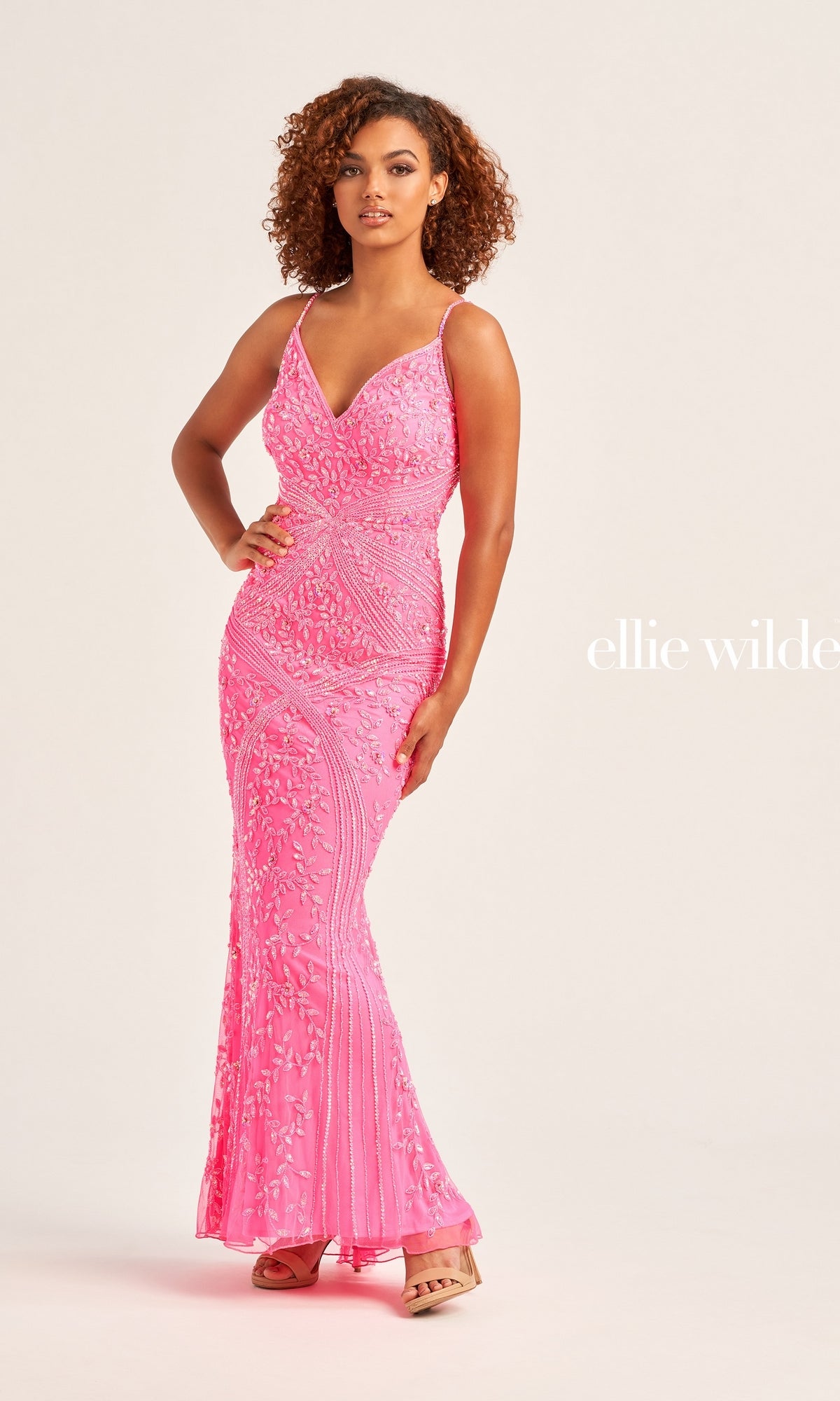 A woman poses in the Long Beaded Ellie Wilde V-Neck Prom Dress EW35065—a fitted, bright pink gown with thin straps, a mermaid silhouette, and shimmering beaded details.